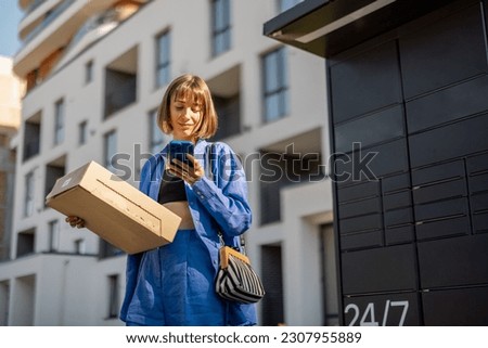 Young woman using smart phone while standing with a parcel delivered with post office machine with automatic lockers. New technologies in delivery service, self picking Royalty-Free Stock Photo #2307955889