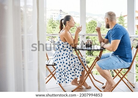 Wife and husband drinking hot beverage in mugs sit on hotel balcony enjoying city view. Married couple spends summer vacation together