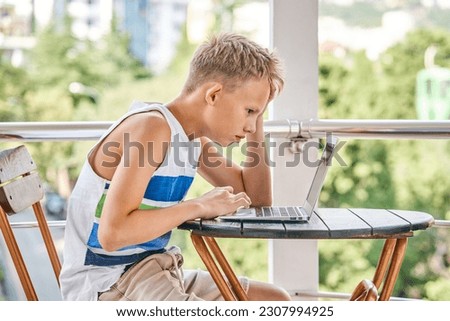 Preteen boy with serious expression sitting at table watches videos on laptop. Schoolboy enjoys watching videos during summer holidays in hotel