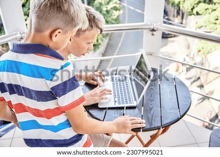 Brother siblings look together at difficult game on grey laptop on hotel balcony. Preteen schoolboy tries to win serious game during summer holidays
