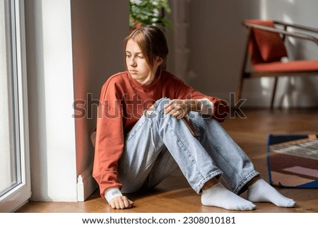 Upset teen girl sit on floor sadly look out window worried about teenage problem at school and communication with parent. Worried girl tensely suffer about bullying at school, unrequited love with boy Royalty-Free Stock Photo #2308010181