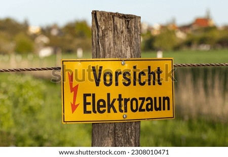Vorsicht Elektrozaun (Electric fence caution). Yellow warning sign on the bar of the electric fence. Blured meadow in the background. Agriculture, fencing, fence for livestock.