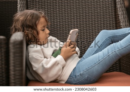 a small, pretty girl, curly brunette, with a smartphone in her hands, lying on a street chair, the concept of gadgets for children