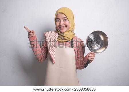 Portrait of cheerful young Asian Muslim woman housewife wearing apron showing frying pan and pointing to copy space isolated on white background.