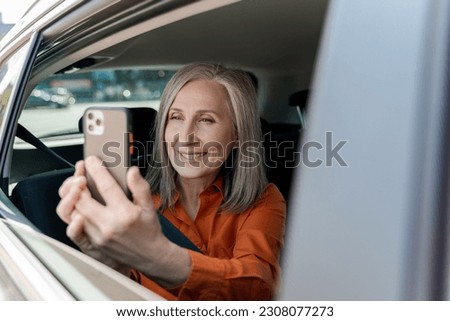 Smiling attractive senior woman holding mobile phone, taking picture looking at window window sitting inside taxi. Travel, vacation, Transportation concept