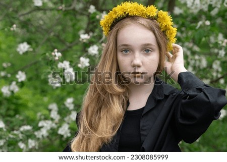 girl of 13 with long blond hair and a wreath of dandelions. A thoughtful blonde in a spring blooming garden looks at the photographer's camera.