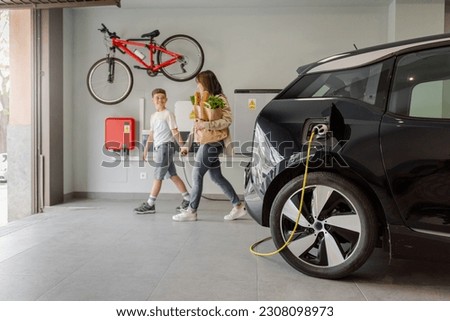 Electric vehicle charging station in private home with happy mother and son walking alongside, blurred family leaving the house, with a bicycle hanging on the wall. Horizontal. Royalty-Free Stock Photo #2308098973
