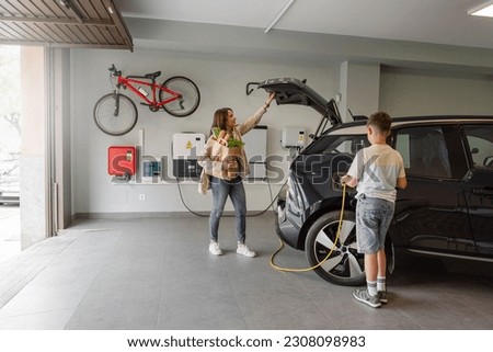 Son plugs EV charger from charging station to electric vehicle in private home while mother takes groceries from the car on daytime. Sustainable Alternative Lifestyle. Horizontal copy-space. Royalty-Free Stock Photo #2308098983