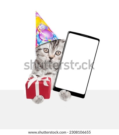 Cute kitten wearing party cap holds gift box and shows big smartphone with blank screen above empty white banner. isolated on white background