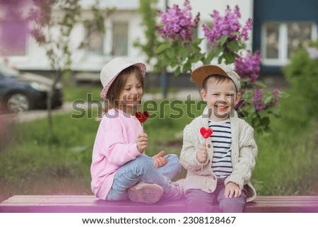 Lifestyle. Little beautiful children are playing on a bench with candy lollipops on a stick in the shape of a heart. The concept of childhood friendship and first love.
