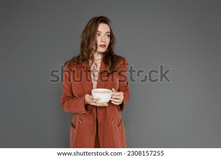 sophisticated young woman with brunette hair wearing brown and trendy suit with blazer and holding cup of coffee while looking away on grey background, work-life balance