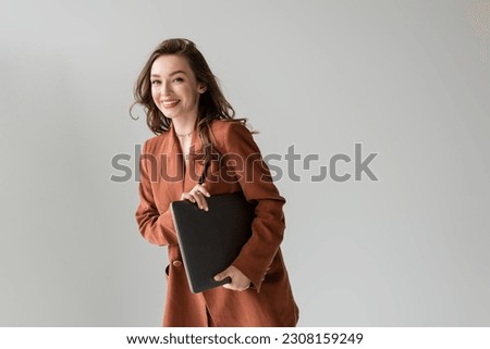 happy young woman with brunette hair and necklace wearing brown and trendy suit with blazer, looking at camera and holding laptop while standing on grey background, freelancer, remote work Royalty-Free Stock Photo #2308159249