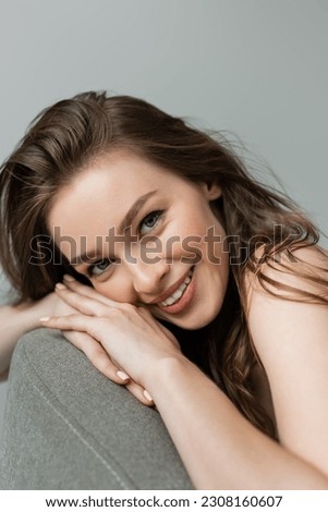 Portrait of smiling and pretty young woman with natural makeup and brunette hair looking at camera while relaxing on comfortable grey armchair isolated on grey
