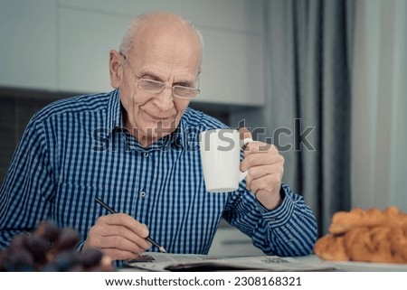 Happy senior man with pen in hand wearing eyeglasses solving crossword and drinking coffee sitting at kitchen table indoor. Royalty-Free Stock Photo #2308168321