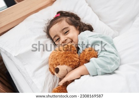 Adorable hispanic girl hugging teddy bear lying on bed at bedroom