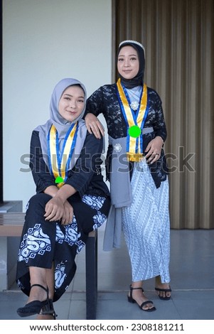 two young women taking pictures together, one sitting and the other standing, commemorating graduation, kebaya clothes