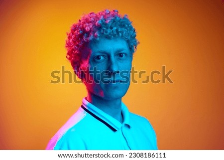 Portrait of mature blonde man with curly hair looking serious at camera against orange studio background in blue neon light. Concept of human emotions, lifestyle, youth, facial expression