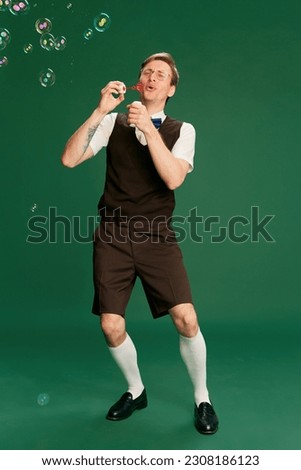 Portrait of man in stylish classical clothes, shorts and vest posing, blowing bubbles over green studio background. Concept of emotions, facial expression, lifestyle, retro fashion. Ad