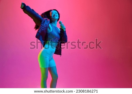 Portrait of happy, smiling, young girl singing in microphone against pink studio background in neon light. Leisure. Concept of emotions, facial expression, youth, lifestyle, inspiration, sales, ad Royalty-Free Stock Photo #2308186271