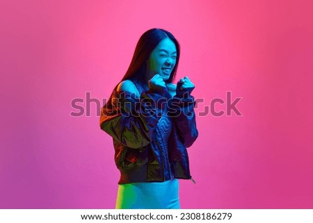 Anger, irritation. Portrait of young korean girl in casual clothes posing with fists up against pink studio background in neon light. Concept of emotions, facial expression, youth, lifestyle, ad