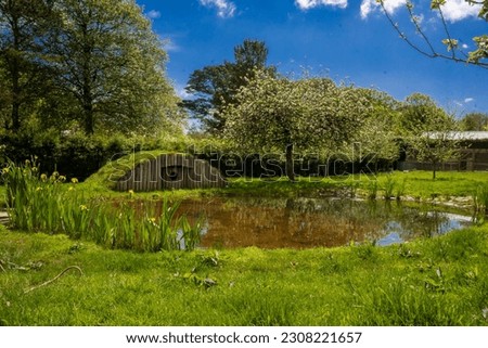 landscape picture of a domed  duck hobbit house over looking a pond surrounded my foliage plants and trees green grass and a blue sky 