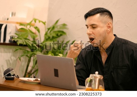Side view of happy handsome young man eating dessert while working on laptop at home