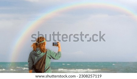 A blogger and hippie girl takes pictures of a rainbow on a mobile phone. Beautiful rainbow over the sea. The concept of travel. Selective attention to the girl. copy space banner