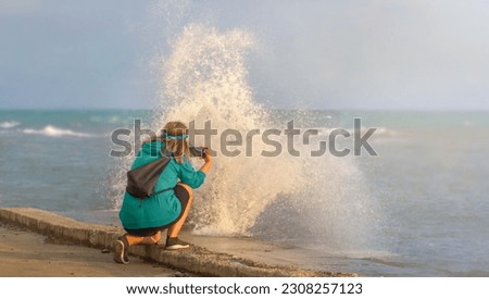 A young hippie blogger girl with curly hair, in a green hoodie on a beach flooded with waves takes pictures of a storm on her phone during sunset. Space for copying.