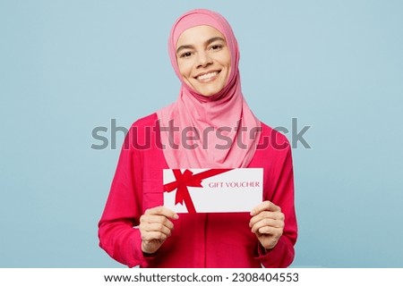 Young fun arabian muslim woman wear pink abaya hijab hold gift certificate coupon voucher card for store isolated on plain pastel light blue cyan background studio. People uae islam religious concept