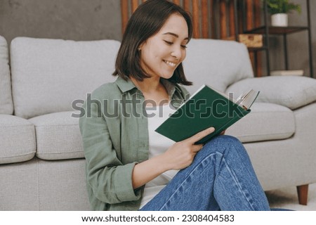 Young smiling smart student woman of Asian ethnicity wears casual clothes reading book novel sits near grey sofa couch stay at home hotel flat rest relax spend free spare time in living room indoor