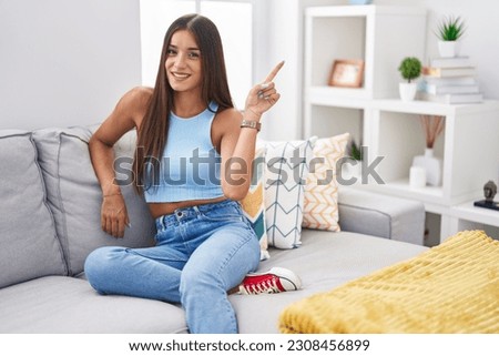 Young brunette woman sitting on the sofa at home with a big smile on face, pointing with hand finger to the side looking at the camera. 