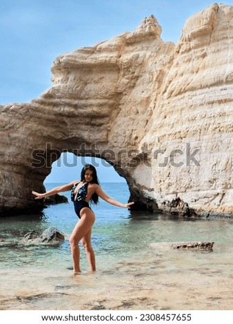 A brunette stands in front of the famous arch in the Sea Caves, white cliffs in the city of Paphos. She is wearing a solid black swimsuit. She poses while standing in the Mediterranean Sea.