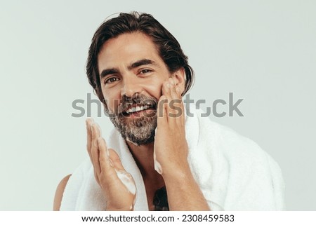 Happy caucasian man takes pride in his grooming routine, carefully applying shaving cream to his beard. With a steady hand and a focused gaze, he prepares his face for a close shave in a studio. Royalty-Free Stock Photo #2308459583