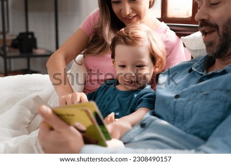 Father reading a bedtime story to his son at home, kid's eye focused on colorful cartoons in book while mother hugs a child. 