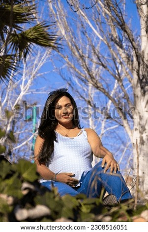Beautiful pregnant Latin woman sitting on grass, smiling, wearing blue clothes and sunglasses. With autumn trees in background
