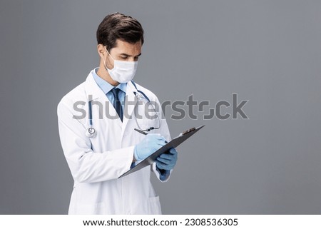 A male doctor in a white coat and a medical mask with a notepad for notes and a patient card in his hand looks at the camera on a gray isolated background, copy space, space for text