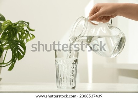 Woman pouring water from jug into glass on white table indoors, closeup Royalty-Free Stock Photo #2308537439