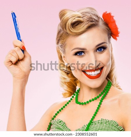 Beautiful young happy smiling woman hold pen, in green pin-up style cloth wear, on rose pink background. Caucasian blond model posing in retro fashion and vintage concept studio photo.