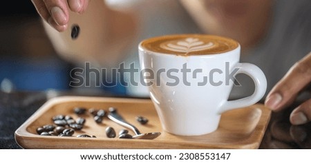 White ceramic cups of cappuccino with latte art Barista make coffee by pouring spills hot milk cream on black coffee. Barista serve holding cup of hot latte and coffee beans on wooden table cafe shop