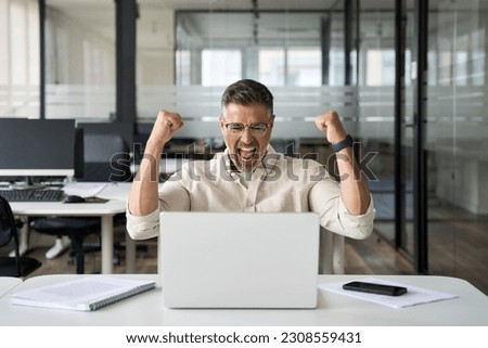 Middle-aged Latin or indian businessman screaming proud cheering with excitement , man read great news on smartphone celebrating victory and success sitting front of laptop computer working at office. Royalty-Free Stock Photo #2308559431