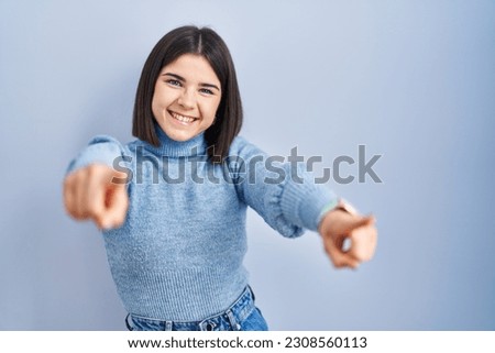 Young hispanic woman standing over blue background pointing to you and the camera with fingers, smiling positive and cheerful 