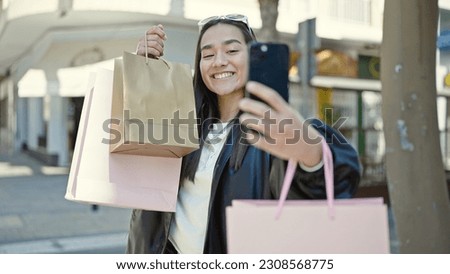 Young beautiful hispanic woman make selfie by smartphone holding shopping bags at street