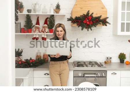 Attractive smiling woman in shirt with laptop at bright kitchen at home. Christmas time