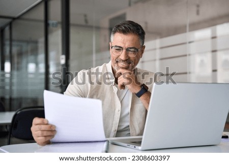 Smiling Latin or Indian male business man accountant analyst holding documents, work at laptop computer doing online trade market tech research. Focused Hispanic businessman with paperwork in office. Royalty-Free Stock Photo #2308603397