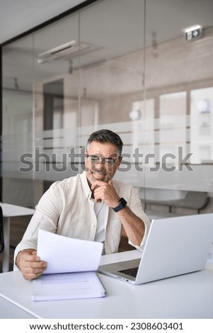 Vertical portrait of Hispanic businessman doing paperwork in office. Happy Latin or Indian male business man holding documents, working at laptop computer doing online trade market tech research. Royalty-Free Stock Photo #2308603401