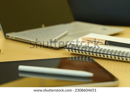 Electronics items on a table along with a notebook, mobile and pen. Picture contains a tablet, a laptop with touch pencil, writing notebook with a white pencil.