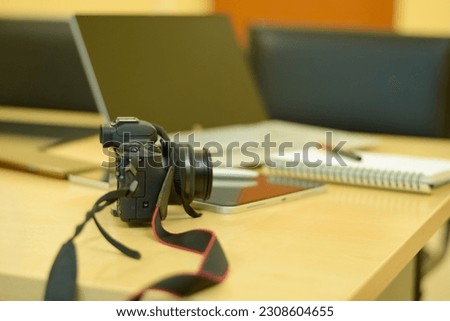 Electronics items on a table along with a notebook, camera, lens, mobile and pen. Picture contains a tablet, a laptop with touch pencil, writing notebook with a white pencil.
