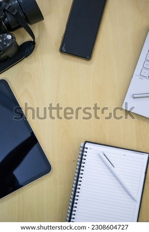 Electronics items on a table along with a notebook, camera, lens, mobile and pen. Picture contains a tablet, a laptop with touch pencil, writing notebook with a white pencil.