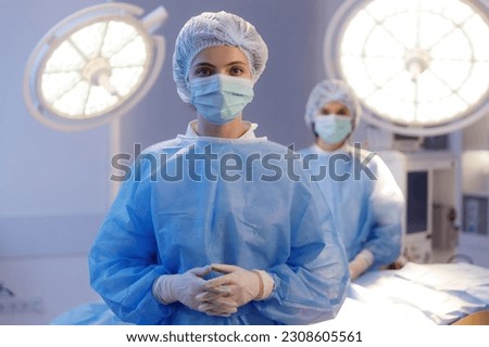 A close up shot of a beautiful nurse wearing scrubs, a medical mask and a medical hairnet standing in a hospital room and behind her another nurse Royalty-Free Stock Photo #2308605561
