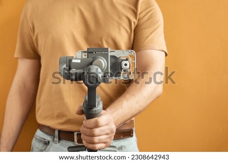 Man filming with a smartphone using a gimbal stabilizer on yellow background. Using three-axis electronic stabilizer to make vlogs or video shooting. Royalty-Free Stock Photo #2308642943